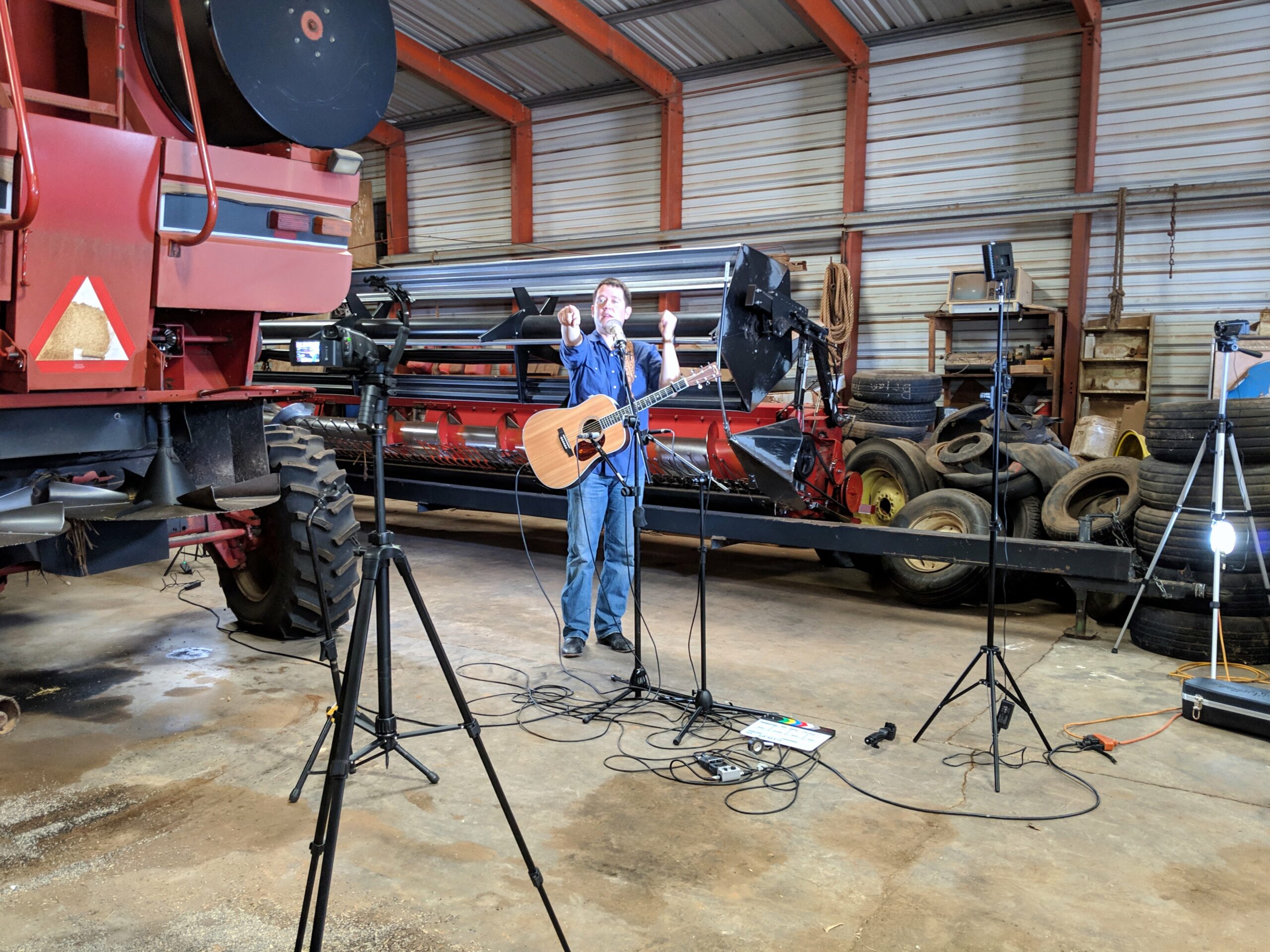 A camera on a tripod filming a musician performing beside a combine harvester in a machine shed